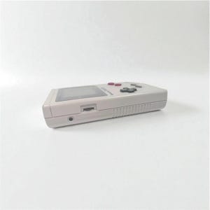 May include: A light gray handheld video game console with a rectangular screen, red and black buttons, and a cartridge slot. The device is shown from a side angle against a white background.