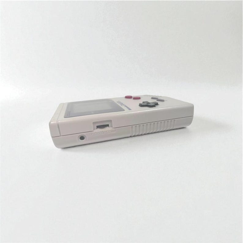 May include: A light gray handheld video game console with a rectangular screen, red and black buttons, and a cartridge slot. The device is shown from a side angle against a white background.