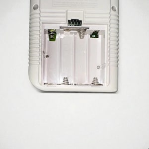 May include: A close-up of the battery compartment of a white handheld gaming device. The compartment is open, revealing four empty battery slots with metal springs. The device has a ridged design on the side.