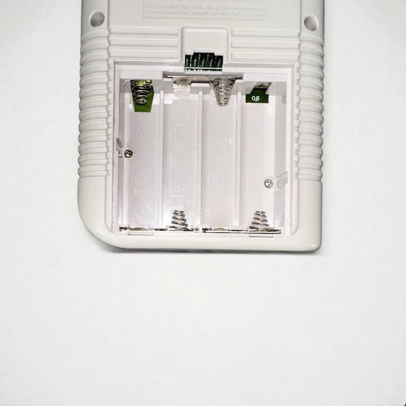 May include: A close-up of the battery compartment of a white handheld gaming device. The compartment is open, revealing four empty battery slots with metal springs. The device has a ridged design on the side.
