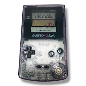May include: A handheld Game Boy Color gaming console with a translucent purple plastic shell. The screen displays the Tetris title screen with the text 1 PLAYER and 2 PLAYER. It features a black directional pad, A and B buttons, and start buttons.