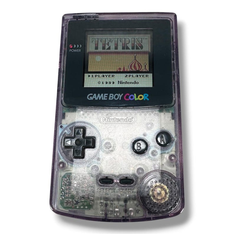 May include: A handheld Game Boy Color gaming console with a translucent purple plastic shell. The screen displays the Tetris title screen with the text 1 PLAYER and 2 PLAYER. It features a black directional pad, A and B buttons, and start buttons.