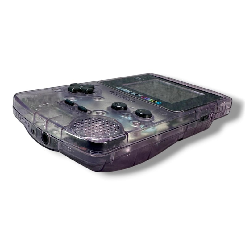 May include: A handheld Game Boy Color gaming console with a translucent purple plastic shell. The device features a central screen, a black directional pad, two black action buttons, a start button, a select button, and a circular speaker grille.