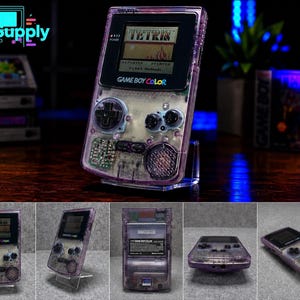 May include: A transparent purple Game Boy Color handheld gaming console featuring a black screen bezel, a directional pad, two action buttons, and a speaker. The device displays the Tetris game title on its screen and includes a battery compartment.