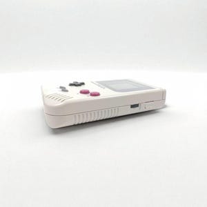 May include: A white handheld gaming console with a black directional pad, pink buttons, and a rectangular screen. The console has a speaker and a power switch on the side. The device is a classic retro gaming console.
