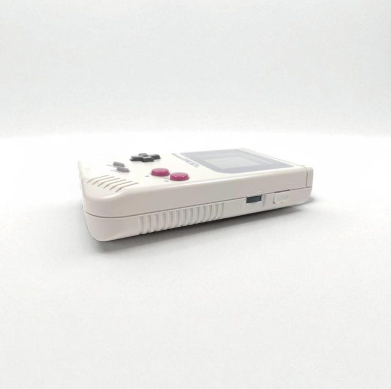 May include: A white handheld gaming console with a black directional pad, pink buttons, and a rectangular screen. The console has a speaker and a power switch on the side. The device is a classic retro gaming console.