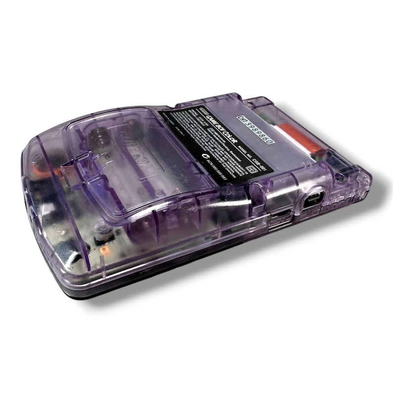 May include: A translucent purple handheld game console with a visible battery compartment and internal circuitry. The back label displays the text GAME BOY COLOR, model number CGB-001, and a serial number CH13989860, with a metallic battery contact.