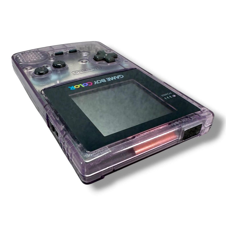 May include: A handheld Game Boy Color gaming console featuring a translucent purple plastic shell. The device includes a black screen bezel with the text GAME BOY COLOR, a dark gray directional pad, two black action buttons, and a power indicator light.