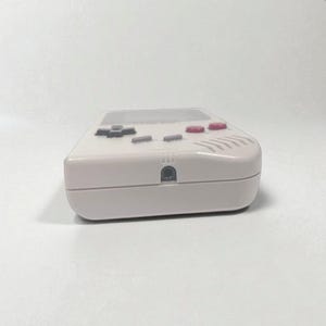 May include: A light gray handheld gaming console with a rectangular design. The console features a directional pad, action buttons, and a screen. The buttons are gray and red. The device has a headphone jack.