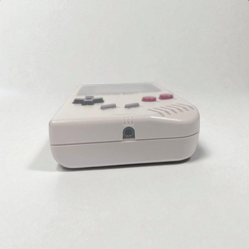 May include: A light gray handheld gaming console with a rectangular design. The console features a directional pad, action buttons, and a screen. The buttons are gray and red. The device has a headphone jack.