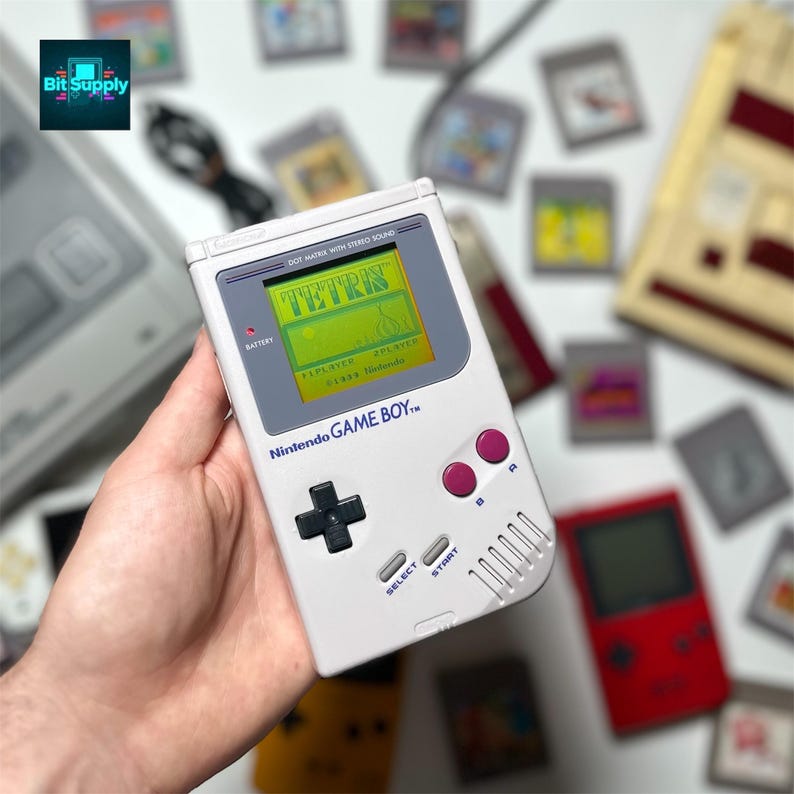 May include: A white Nintendo Game Boy handheld console displaying the game Tetris on its screen. The console has a black directional pad, pink buttons, and the text "Nintendo GAME BOY" printed on it. Several game cartridges are scattered around the console.