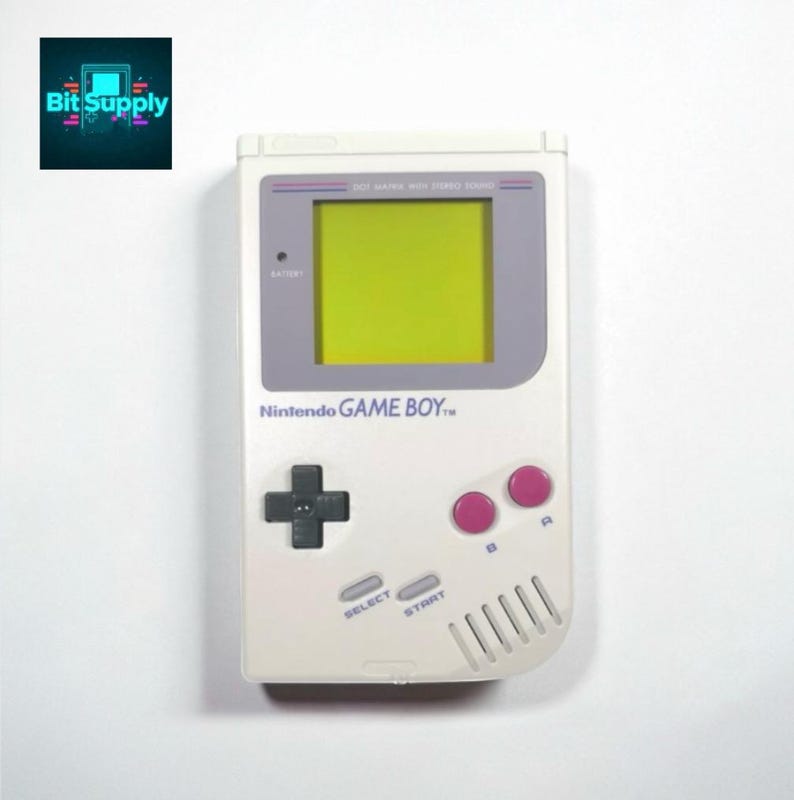 May include: A white Nintendo Game Boy handheld console with a green screen, gray accents, and pink buttons. The console features a directional pad, select and start buttons, and the text "Nintendo GAME BOY". The Bit Supply logo is in the upper left corner.
