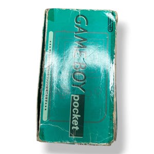 May include: A rectangular teal cardboard box for a Nintendo Game Boy Pocket handheld gaming console. The front features the text GAME BOY pocket in white and black, a Nintendo logo, and Japanese text along the left side. The box shows visible wear.