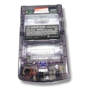 May include: A translucent purple handheld video game console with a visible internal circuit board. The back features a black label with white text reading Nintendo Game Boy Color, Model No. CGB-001, and a battery compartment with Game Boy embossed.