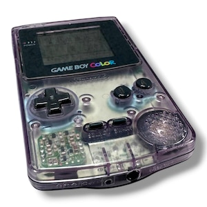 May include: A handheld Game Boy Color gaming console featuring a translucent purple plastic shell. The device includes a black screen bezel with the text GAME BOY COLOR, a black directional pad, two black action buttons, and two small black menu buttons.