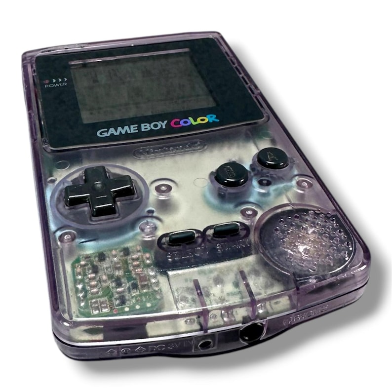 May include: A handheld Game Boy Color gaming console featuring a translucent purple plastic shell. The device includes a black screen bezel with the text GAME BOY COLOR, a black directional pad, two black action buttons, and two small black menu buttons.
