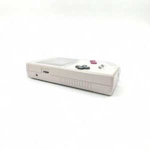 May include: A light gray handheld video game console with a rectangular screen and a set of black and pink buttons. The console has a directional pad and a speaker. The device is set against a white background.