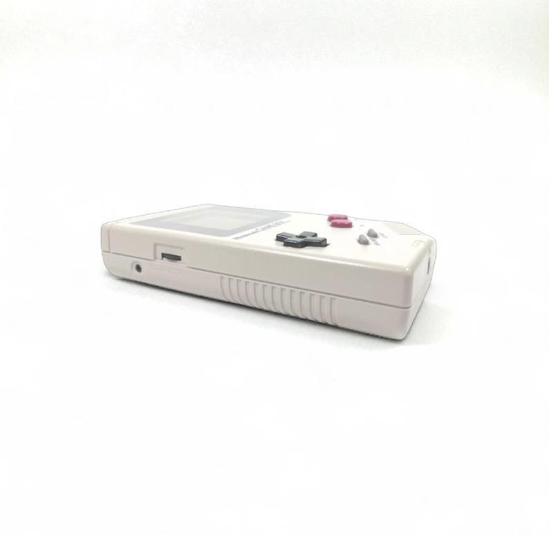 May include: A light gray handheld video game console with a rectangular screen and a set of black and pink buttons. The console has a directional pad and a speaker. The device is set against a white background.