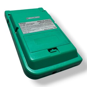 May include: A green handheld video game console, model MGB-001, viewed from the back. The device features a battery compartment cover and a rectangular label with technical specifications, including the text Nintendo 1989-1996 and Made in China.