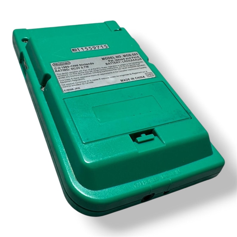 May include: A green handheld video game console, model MGB-001, viewed from the back. The device features a battery compartment cover and a rectangular label with technical specifications, including the text Nintendo 1989-1996 and Made in China.