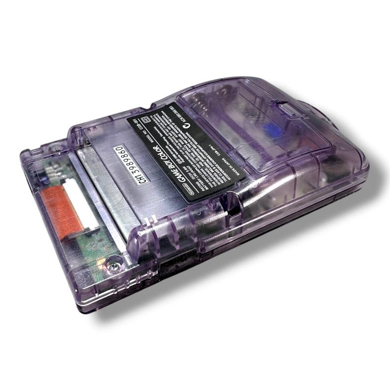 May include: A translucent purple handheld game console with a visible internal battery compartment and circuit board. The back features a black label with white text reading GAME BOY COLOR, model number CGB-001, and serial number CH13989860.
