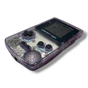 May include: A handheld Game Boy Color gaming console featuring a translucent purple plastic shell. The device includes a black screen bezel with the text GAME BOY COLOR, a directional pad, A and B buttons, Start and Select buttons, and a speaker.