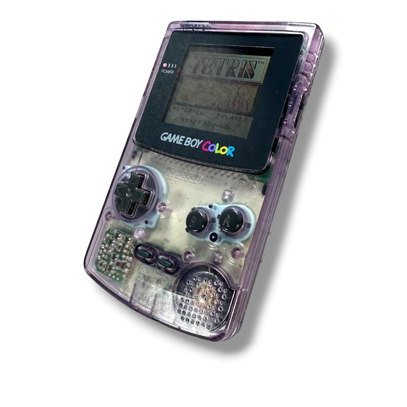 May include: A handheld Game Boy Color gaming console with a translucent purple plastic shell. The device features a black screen bezel displaying the Tetris game title, a directional pad, A and B buttons, a speaker grille, and internal components.