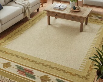 May include: A rectangular beige area rug with a geometric border in shades of green, red, and brown. The rug has a fringed edge and is placed in a living room setting with furniture and plants.