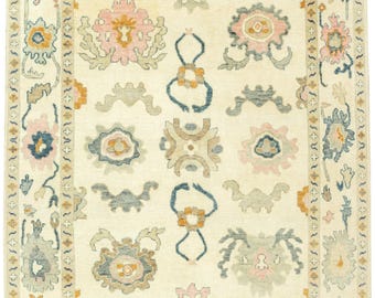 May include: Cream-colored area rug with a floral pattern in shades of pink, blue, and gold. The rug has a border with repeating floral and geometric designs. The central field features larger floral motifs and stylized shapes.