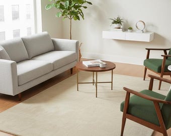 May include: A modern living room with a light gray sofa, two green armchairs, and a round wooden coffee table. A beige rug covers the floor, and a large window lets in natural light. A potted plant adds a touch of greenery.