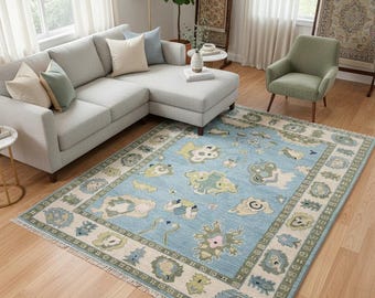 Puede incluir: Una alfombra rectangular grande con un campo azul claro y un borde crema. La alfombra presenta un patrón repetitivo de motivos florales y de nubes estilizados en tonos verdes, amarillos y rosas. Está colocada sobre un suelo de madera en un salón.