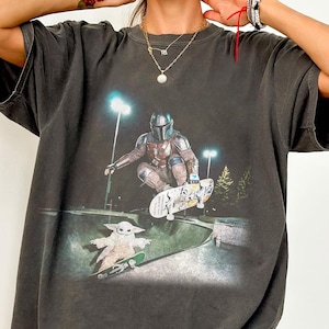 May include: Dark gray Comfort Colors t-shirt featuring a graphic of The Mandalorian and Grogu skateboarding in a skate park. The shirt has a distressed look and the text "Comfort Colors Pepper" is printed on the bottom right.