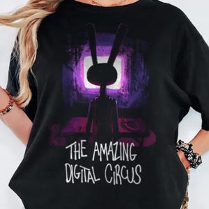 May include: A black short-sleeved t-shirt featuring a graphic print of a rabbit-like character standing before a glowing purple screen. Below the character, the text reads THE AMAZING DIGITAL CIRCUS in a white, hand-drawn style font.