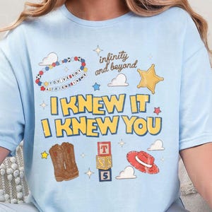 Pode incluir: Uma t-shirt azul-clara com uma estampa gr&aacute;fica com o texto "I KNEW IT I KNEW YOU", "infinity and beyond" e "YOU'VE GOT A FRIEND IN ME". O design inclui ilustra&ccedil;&otilde;es de uma pulseira de contas, uma estrela, nuvens, botas de cowboy, blocos e um chap&eacute;u.