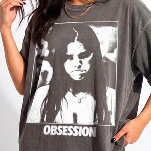 May include: A charcoal gray short-sleeve t-shirt featuring a black and white graphic print of a person with long hair. The word OBSESSION is printed in white capital letters below the image. The shirt has a crew neckline and a relaxed fit.