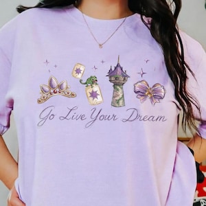 May include: A light purple t-shirt with a graphic design featuring a crown, cards, a tower, and a bow. The text "Go Live Your Dream" is written in cursive. The shirt also has the words "Comfort Colors" and "Orchid" printed on it.