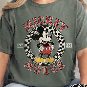 May include: Olive green t-shirt featuring a vintage Mickey Mouse graphic. The design includes the words "Mickey Mouse" in red, a checkered circle, and the year "1928." The shirt also has the words "Comfort Colors" and "Moss" in a white oval.
