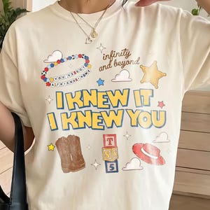 May include: An ivory short-sleeve t-shirt featuring a graphic print with the text "I KNEW IT I KNEW YOU", "infinity and beyond", and "YOU'VE GOT A FRIEND IN ME". The design includes a beaded bracelet, a star, cowboy boots, blocks, and a hat.