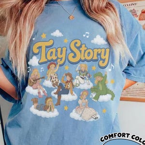 May include: A light blue t-shirt featuring the text Tay Story in yellow letters. The graphic includes multiple illustrations of a blonde character in various outfits, including a cowgirl, a dinosaur costume, and a dress, all positioned on white clouds.