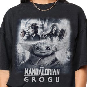 May include: Black t-shirt with a graphic of Grogu from The Mandalorian series. The design includes characters like Luke Skywalker and The Mandalorian. The text "THE MANDALORIAN and GROGU" is printed below the image.