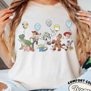 May include: Cream-coloured t-shirt with a vibrant graphic of Toy Story characters and balloons. The design includes Woody, Buzz Lightyear, and other characters from the animated film. The shirt has the words "Comfort Colors" and "Ivory" printed on it.