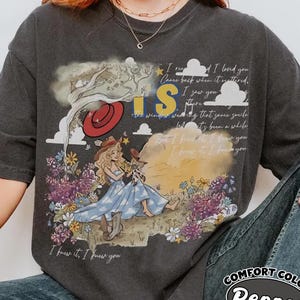 May include: A charcoal gray short-sleeve t-shirt featuring a graphic print of a person with long blonde hair sitting in a field of purple and yellow flowers. The design includes a red hat, clouds, a tree, and the text "I knew it, I knew you."