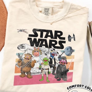 May include: Ivory-coloured t-shirt featuring a graphic of Muppets characters as Star Wars characters. The words "STAR WARS" are printed in black. The shirt has a crew neck and a Comfort Colors tag.