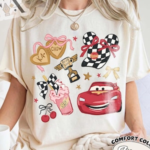 May include: Ivory-colored t-shirt featuring cartoon graphics of a red race car, checkered flags, and heart-shaped cookies. The shirt also includes a milkshake, cherries, and the text "Piston Cup". The shirt is labeled "Comfort Colors Ivory".