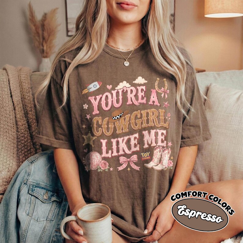 May include: A dark brown short-sleeve t-shirt featuring the pink text "YOU'RE A COWGIRL LIKE ME" with graphic illustrations of a rocket, a cowboy hat, a bow, stars, and a pair of cowboy boots. The shirt has a vintage-style washed appearance.