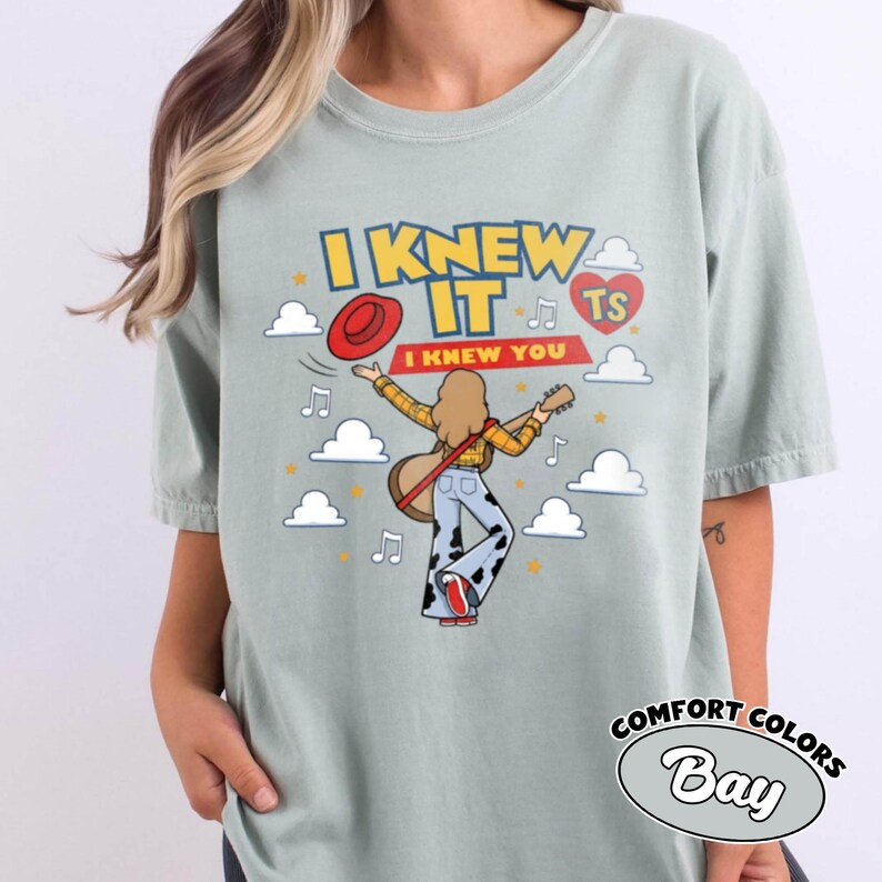 May include: A light sage green t-shirt featuring a graphic of a person with long hair playing a guitar, wearing a yellow plaid shirt and cow-print trousers. The text reads I KNEW IT and I KNEW YOU, accompanied by a red hat, musical notes, and clouds.