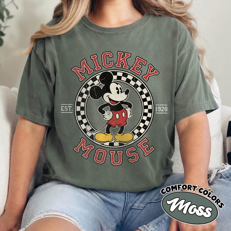 May include: Olive green t-shirt featuring a vintage Mickey Mouse graphic. The design includes the words "Mickey Mouse" in red, a checkered circle, and the year "1928." The shirt also has the words "Comfort Colors" and "Moss" in a white oval.