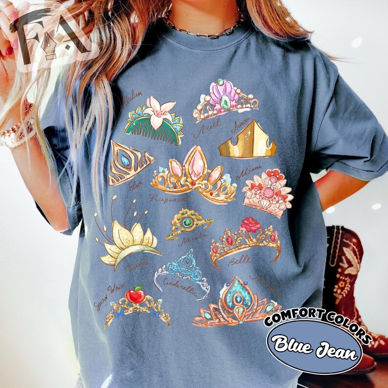 May include: Blue jean Comfort Colors t-shirt featuring colorful illustrations of various tiaras. The tiaras are labeled with names like Ariel, Belle, and Cinderella. The shirt has a relaxed fit and is made of soft material.