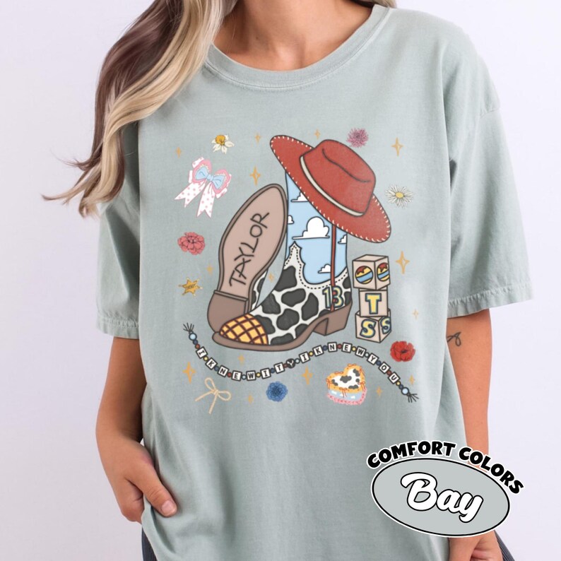Op de afbeelding: Een saliegroen t-shirt met een grafische print van een cowboylaars met koeienprint, een rode hoed, blokken met de letters T, S en het getal 13, een strik en een kralensnoer met de tekst I KNEW IT WHEN YOU.