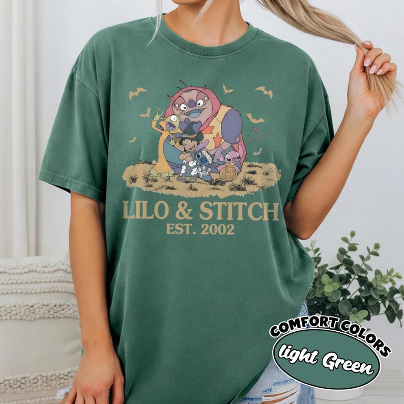 K&ouml;nnte beinhalten: Hellgr&uuml;nes T-Shirt mit einer Grafik von Lilo & Stitch-Charakteren. Das Design enth&auml;lt den Text "LILO & STITCH EST. 2002" im Vintage-Stil. Das Shirt hat auch ein "COMFORT COLORS light Green"-Label.